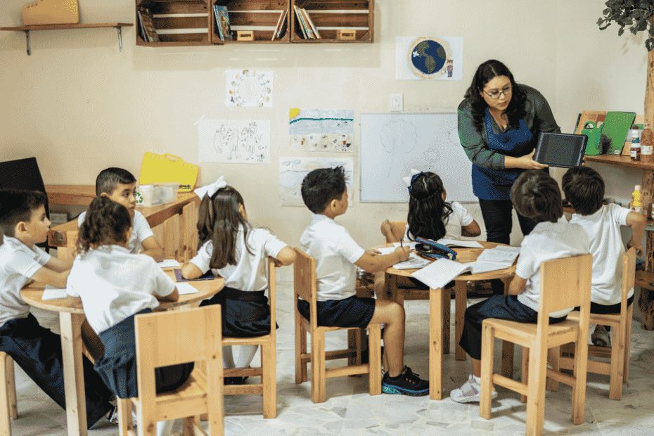 A teacher showing a tablet to a group of young students sitting at wooden tables in a classroom.